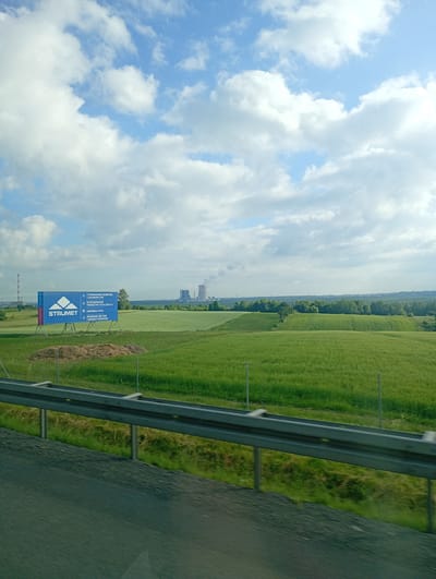 The image depicts a rural landscape in Mysłowice, Poland, on a sunny day with scattered clouds. In the mid-ground, a large blue billboard for "STRUMET" is prominently displayed. The billboard features a white logo and text in Polish, indicating services such as galvanizing, welding, and steel fabrication. Behind the billboard, a vast green field stretches towards a treeline. In the far distance, a power plant with cooling towers and a tall smokestack is visible, with some emitting steam or smoke. A highway guardrail runs across the foreground of the image, obscuring the lower portion of the scene. The overall impression is one of an industrial area juxtaposed with agricultural land.