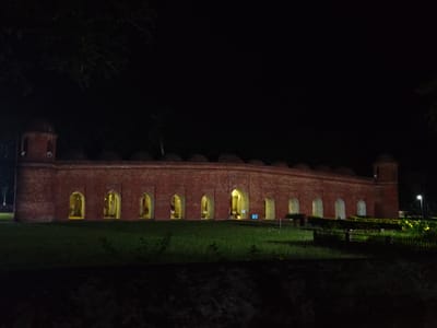 The image shows the exterior of a long, red brick building with multiple arched openings, illuminated by internal lighting and external spotlights, at night. The building features domed structures at either end and appears to be a historical site. In front of the building is a well-maintained lawn with some small plants and a low wooden fence. To the right, a street lamp is visible, along with some yellow flowers and a larger green bush. The background is dark, indicating nighttime, with the faint silhouette of trees. This is the exterior of the Khan Jahan Ali's mosque, located in Bagerhat, Bangladesh.
