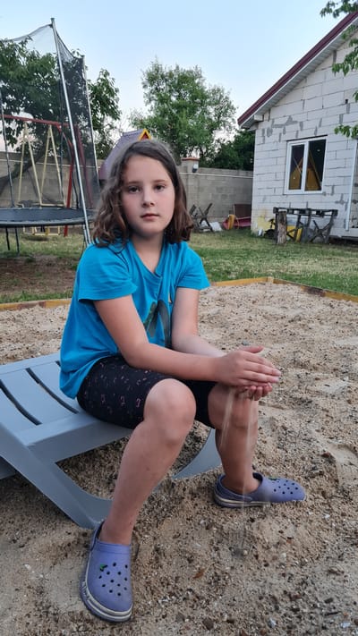 This media file depicts a young girl sitting in a sandbox, playing with sand. She is wearing a blue t-shirt, black shorts with a pink pattern, and light purple Crocs. Her hair is brown and wavy. The girl is holding sand in her hands, letting it sift through her fingers.
The scene appears to be a backyard. In the background, there is a partially constructed house with white cinder blocks and a red-trimmed roof. Adjacent to the house, there is a green lawn and a trampoline with a safety net. The sandbox itself is a wooden-framed area filled with sand, and the girl is seated on a gray lounge chair.
The lighting suggests it is either early morning or late afternoon, with a soft, diffused light. The sky is a pale blue, indicating fair weather. The overall mood is relaxed and casual, typical of a child playing outdoors. The location context provided places this scene in Bilhorod-Dnistrovs'kyi, Ukraine. There is no visible text in the image.