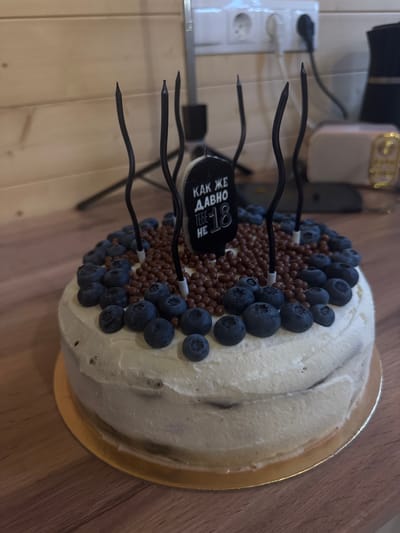 This is a close-up, slightly overhead shot of a cake decorated with blueberries and chocolate sprinkles. The cake is on a golden cake board, placed on a wooden surface, likely a table. The cake is covered in white frosting and has a rustic, layered appearance. Several black, curved candles are inserted into the cake, pointing upwards. A small, black tag with white text is also attached to one of the candles. The text on the tag is in Russian and appears to say "КАК ЖЕ ДАВНО ТЕБЕ НЕ" with a number "18" prominently displayed. This suggests the cake is for an 18th birthday celebration. The lighting in the image is somewhat dim, indicating it might be indoors or late in the day. The background shows a wall with horizontal wooden planks and some electrical outlets, further suggesting an indoor setting. No people are visible in the image. The overall mood is festive, likely related to a birthday event.