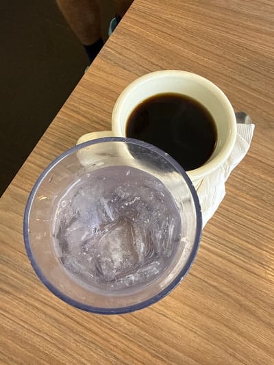 The image shows a top-down view of a wooden table with two drinks. On the left is a white mug filled with dark liquid, likely coffee. The mug is placed on a white napkin. To the right of the mug is a clear plastic cup filled with ice and a clear liquid, possibly water or a soda. The cup has a blue rim. The scene appears to be indoors, possibly a restaurant or cafe, given the presence of a table and drinks. The lighting suggests it could be daytime. There is no visible text in the image. The overall impression is a casual setting, possibly a moment of rest or a meal.