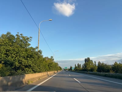 The image shows a view from a vehicle driving on a highway in the city of Changilari, Georgia. The highway has multiple lanes and is bordered by concrete barriers and lush green trees. The sky is clear blue with a single white cloud. There are several streetlights along the highway, and a few cars are visible in the distance. Green signs with white text are placed along the roadside, indicating directions to cities. The sun appears to be low in the sky, casting long shadows on the road.