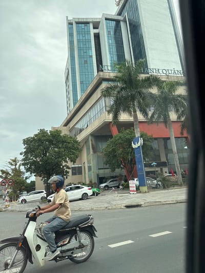 A man rides a motorcycle on a street in Nam Đông Hà Ward, Vietnam. The man, wearing a light brown t-shirt and blue pants, has a blue helmet and is looking forward. His motorcycle is dark brown and white. In the background, a tall, modern building with blue-tinted glass windows stands against a cloudy sky. Palm trees and other green foliage are visible in front of the building. Several cars, including white sedans and a silver SUV, are parked or driving on the street. Traffic signs and a blue directional sign are also present.