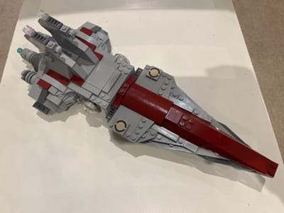 A LEGO model of a spacecraft, primarily light grey with dark red and dark grey accents, rests on a flat, white surface. The model is elongated and wedge-shaped, with a broader stern featuring multiple engine nozzles, including three prominent grey ones and a smaller light blue circular element. Four fin-like structures with transparent clear tips are visible at the stern. Dark red plating forms a central spine and tapers to a point at the bow. Two circular flat tiles with light tan inner rings are visible on the model's mid-section. The white surface shows minor dark specks, possibly dust or debris. In the background, a light beige or cream-colored textured surface, possibly carpet or flooring, is partially visible. Shadows are cast beneath and to the right of the LEGO model. No people or actions are depicted. The specific location in the city of Abbotsbury, United Kingdom, cannot be determined from the visual information provided in the image.
