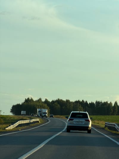 In Togashevo, Russia, a silver Volkswagen Tiguan with license plate A766MC799 drives on a multi-lane highway under a pale sky. The car is positioned in the right lane, facing away from the camera. The highway has a white dashed line marking the lanes and a solid white line on the right edge. Guardrails line the side of the road. In the distance, several trucks and other vehicles are visible, as well as a dense line of trees and green fields. The lighting suggests it is late afternoon or early evening.