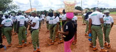 The image displays a group of approximately 25-30 young adults gathered outdoors in the city of Peyer, Nigeria. The majority are uniformly dressed in white short-sleeved t-shirts printed with "NYSC" in green, green camouflage cargo pants, green baseball caps, and tan ankle boots. They are standing on reddish-brown, uneven soil under an overcast sky. In the mid-ground, a female individual, seen from behind, wears a black t-shirt, pink trousers, and a pink headwrap, and balances a clear plastic bucket filled with pale, spherical fruits on her head. The uniformed individuals are standing in various orientations, some appearing to converse, others standing idly; one male in the foreground holds a blue and orange water bottle. The background reveals several single-story buildings featuring orange-brown pitched roofs, interspersed with green trees and vegetation.