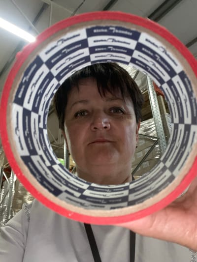 In a warehouse environment, a woman is holding a roll of red tape in front of her face, creating a framing effect. She has short, dark hair and is looking directly at the camera. The tape has a distinctive blue and white checkered pattern with repeated text. Behind her, metal shelves are visible, stocked with cardboard boxes. The lighting suggests overhead fluorescent lights, typical of an industrial setting. The location is identified as Терволово, Russia.