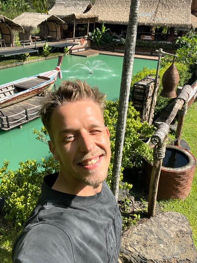 The media file is a portrait-oriented selfie taken outdoors in a tropical setting. The subject is a man with short blonde hair and a beard, smiling at the camera. He is wearing a dark grey t-shirt. The background suggests a resort or tourist attraction with thatched-roof buildings and a vibrant green body of water, possibly a pool or a canal. A traditional wooden boat with colorful stripes is visible on the water.
In the upper right of the frame, there is a large, rustic clay pot with a dark interior, placed on a wooden structure. Woven baskets hang from wooden beams, and there are stacks of wood. Lush green foliage surrounds the area. The sunlight suggests it is daytime, and the weather appears to be sunny and warm. The location is identified as Tambon Pa Kae Bo Hin, Thailand, which aligns with the visual cues of tropical vegetation and traditional architecture. A small fountain is visible in the water, adding to the scenic ambiance. There is a red life preserver hanging on a railing near one of the buildings. Text is visible on a sign in the upper left portion of the image, but it is difficult to read clearly. The overall mood is relaxed and happy, reflecting the subject's smile and the pleasant environment.