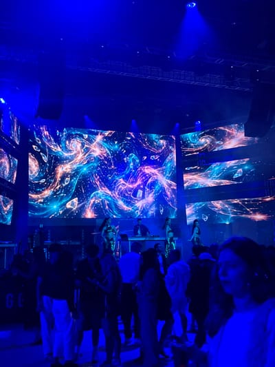 The image captures a vibrant nightclub scene illuminated by an intense blue light. A large LED screen at the back displays a swirling, abstract pattern of blues, pinks, and oranges, creating a dynamic visual backdrop. On stage, a DJ is visible behind a console, with two dancers in sparkly outfits positioned on either side. In the foreground and mid-ground, a crowd of people are gathered, some standing and talking, others seemingly dancing or observing the stage. The environment appears to be a large indoor venue, likely a club or concert hall, with elevated structures and lighting rigs visible towards the ceiling. The overall impression is one of an energetic and lively event, set against a backdrop of impressive visual effects. There is no indication that this scene is located in Akhalubani, Georgia.