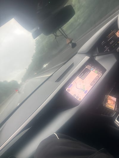 The image depicts the interior of an Audi e-tron vehicle, looking forward through the windshield, taken at 19:21 in the early evening during overcast and possibly rainy weather. The vehicle is located on a road within the city of Uttlesford, United Kingdom, specifically on or approaching the M11 motorway near Stansted Airport, heading towards Colchester.
The dashboard features an infotainment screen displaying a navigation map indicating a remaining distance of 3.1 miles, an estimated arrival time of 19:33 (12 minutes travel time), and a destination of "Colchester, p. Stansted." The map shows the vehicle's position near Thremhall Avenue, an access road to Stansted Airport. The vehicle's gear selector is in 'D' (drive), and a temperature reading of '2.9' is visible. A rearview mirror is present at the top, from which a decorative air freshener, composed of a wooden prism and a dark geometric object, hangs.
To the right of the main infotainment screen, a secondary smartphone is visible, displaying a media player interface featuring the face of Greek singer Leuteris Pantazis, confirming the music playing which is also shown on the main screen. The lower right portion of the image shows dark clothing with a white stripe, suggesting the presence of a person, likely the driver.
Through the windshield, a wet, multi-lane road or motorway is visible, lined with trees and greenery. Red taillights of other vehicles ahead are faintly discernible, indicating traffic flow. The sky appears bright but diffused, consistent with overcast conditions.