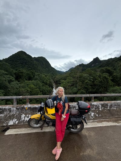 A woman with blonde hair stands next to a yellow motorcycle on a bridge. She is wearing a dark blue t-shirt, red pants, and pink Crocs. The motorcycle has a black helmet with a yellow visor and a black storage box on the back. The bridge has a concrete barrier with graffiti. In the background, there are lush green mountains and a river. The sky is overcast. The location is on a bridge in Thượng Trạch Commune, Vietnam.