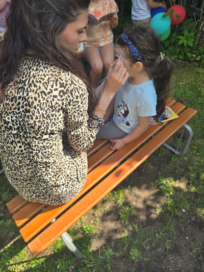 The scene is outdoors, likely a park or garden in Zirndorf, Germany, given the presence of grass, trees, and a wooden bench. A woman with long, dark hair and wearing a leopard-print dress is sitting on the bench. She is face-painting a young girl with dark hair and a blue headband, who is wearing a blue t-shirt with a cartoon character on it and grey pants. The girl is sitting beside the woman on the bench. Another child is partially visible to the left of the girl, wearing an orange and white patterned shirt, with balloons next to them. A colorful book or magazine is also on the bench. The scene appears to be a relaxed, familial activity, possibly at a party or gathering. The lighting is bright and sunny.