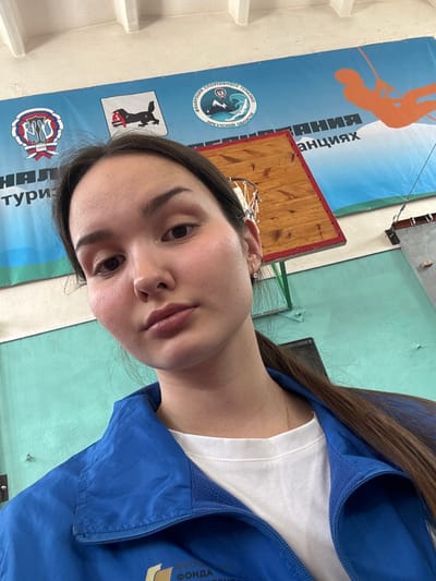 The image displays a young adult female in a close-up selfie within a sports facility in Tayshet, Irkutsk Oblast, Russia. The female has dark brown hair, dark eyes, and a neutral expression, looking directly towards the camera. She is wearing a white t-shirt underneath a blue zippered jacket, with a thin gold chain visible around her neck and two small hoop earrings in her left ear.
The background features a large blue banner stretched across the upper wall. This banner displays multiple logos and text. On the left, there is a red, white, and blue circular emblem featuring stylized Cyrillic initials (likely related to sports tourism) and the number "1895." Next to it is a white shield with a black sable-like animal holding a red ribbon, followed by the text "ФЕДЕРАЦИЯ СПОРТИВНОГО ТУРИЗМА ИРКУТСКОЙ ОБЛАСТИ" (Federation of Sports Tourism of Irkutsk Oblast). A circular emblem depicting mountains and a wave is also present. The banner text partially visible includes "НАЦИОНАЛЬНЫЙ ТУРИЗМ" (National Tourism) on the left and a word fragment ending in "АНЦИЯХ" on the right. To the far right of the banner, an orange silhouette of a person rappelling is depicted against a white and blue background.
Behind the subject's head, a wooden basketball backboard with a red frame and a metal hoop with a white net is visible. Further to the right, a light-colored wall features metallic fixtures, possibly associated with a climbing wall or other sports equipment. The lower section of the visible wall is painted light teal.