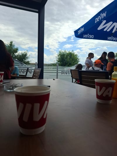 The image shows a cloudy sky over an outdoor seating area at what appears to be a cafe or restaurant. In the foreground, a wooden table is set with two red and white paper cups, one in focus and the other slightly out of focus. An ashtray with cigarettes is also visible on the table, along with a cell phone. In the mid-ground, several people are seated or standing, some under a blue umbrella with "DEVIN" branding. Behind them, a glass barrier separates the seating area from an outdoor space with trees and a parked car. The overall setting suggests a casual gathering in a public outdoor space. This location is identified as being in the city of Drazhevo, Bulgaria.