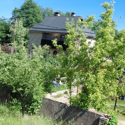 This is a medium-angle, outdoor shot of a two-story brick house with a dark grey, tiled roof and two chimneys, nestled amongst lush green trees under a clear blue sky. The house appears to be in a rural or suburban setting, with a stone retaining wall and a gravel path visible. The foliage is dense and vibrant, suggesting it is either spring or summer. The lighting indicates it is daytime, likely sunny, with shadows cast by the trees and the retaining wall. There is a dark, rectangular outdoor light fixture attached to a wooden pole in the foreground. The overall impression is one of a peaceful, green environment. No people are visible, and no text can be deciphered.