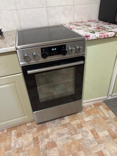 The image shows a close-up view of a modern stainless steel oven in a kitchen. The oven has a glass door, a digital display showing the time "19:13", and several control knobs. Above the oven is a black ceramic cooktop. The kitchen countertops are visible, one with a floral patterned cover. The flooring is a tiled pattern of beige and brown rectangles. To the left of the oven are light beige cabinet doors. The wall behind the oven is tiled with white squares, and to the right, a light green painted wall is visible. The overall scene depicts a domestic kitchen setting. This image is located in Pohreby, Ukraine.