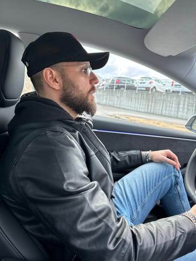 A male individual is seated in the driver's position of a vehicle within the city of Ålesund, Norway. He is observed in profile, facing forward and slightly to the right. The subject has a beard and wears rimless eyeglasses, a black mesh-back baseball cap with a red logo, a black leather jacket over a black hooded garment, and light blue jeans. Black over-ear headphones are positioned around his neck. His left wrist is adorned with multiple metallic bracelets, and a metallic watch or bracelet is on his right wrist. The vehicle's interior features a dark dashboard with a blue ambient light strip, a light-colored headliner, and a visible portion of a dark steering wheel. A panoramic glass roof is partially visible overhead. Through the vehicle's window, an exterior scene depicts an overcast sky above a concrete retaining wall, beyond which numerous vehicles are parked in what appears to be a multi-level parking or storage facility.
