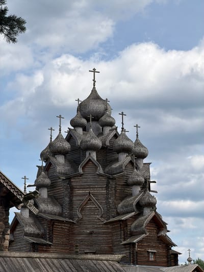 This is a full-frame, eye-level, outdoor, close-up shot of the wooden Church of the Transfiguration in Nevskii Parkleskhoz, Russia. The church has multiple onion domes, each topped with a cross. The structure is made of dark wood, and the domes are covered in wooden shingles. The sky is filled with dramatic, overcast clouds, with some patches of blue sky visible. The top of a pine tree is visible in the upper left corner of the image. The image is focused on the church's intricate wooden architecture. There are no people or animals visible in the frame.