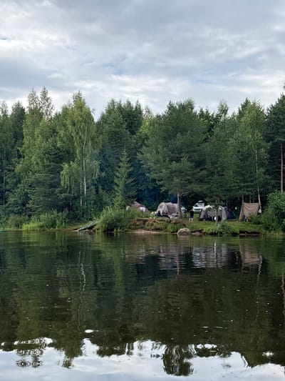 In Anteplevo (Okhotinskoe S/P), Russia, a lakeside campsite is depicted under a cloudy sky. Several tents are pitched on the shore, with a white SUV parked nearby. Some individuals are visible near the tents, suggesting a gathering or recreational activity. The foreground is dominated by a dark body of water, likely a lake, reflecting the trees and sky from the opposite bank. The environment appears to be a wooded area, with dense green foliage bordering the water.
