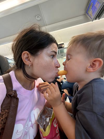 A woman and a young child are on what appears to be public transportation, possibly a train or airplane, with overhead compartments and seating visible. They are positioned close together, facing each other. The woman, wearing a pink t-shirt and a brown shoulder bag, is looking at the child. The child, wearing a grey shirt, is reaching out a snack to the woman, who is also reaching out to take it. Their faces are very close, with the snack between their mouths. A bag of snacks is visible in the foreground. The overall scene captures a moment of playful interaction between the two. The location is identified as Tarragona, Spain.