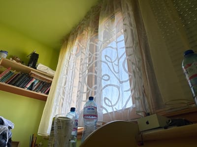 This image is presented sideways and upside down, showcasing an indoor scene, likely a room in a residence located in Topoli, Bulgaria. A large window, covered by two layers of sheer curtains, dominates the center. The inner curtain features an intricate pattern of swirling, abstract lines, while the outer layer is a plain, lighter fabric. Bright daylight filters through the window, indicating it is daytime.

Several plastic water bottles, some with visible condensation, are positioned upside down near the top of the window area. Two prominent bottles with blue caps are labeled "DEVIN", a known Bulgarian mineral water brand. To the right, a wooden shelf is densely packed with books, many featuring titles in Cyrillic script. Notable titles identified through OCR include "ПЕТДЕСЕТ НЮАНСА СИВ" (Fifty Shades of Grey), "МАРТИН СБЛЪСЪК КРАЛЕ" (Martin Clash of Kings), and "МАРТИН ВИХЪР МЕЧОВЕ" (Martin Storm of Swords), suggesting fantasy literature.

Below the bookshelf, a small, light-brown stuffed animal is partially visible, while a pile of clothes rests on top of the shelf. In the upper left, a white box adorned with a black Apple logo is tucked under a surface, with wires connected nearby. The surrounding walls are painted a distinct light, yellowish-green hue. The scene is static, devoid of human presence or ongoing activity, depicting a common, functional interior space.