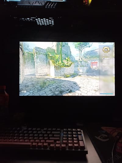 A computer screen displays a video game. The game appears to be a first-person shooter set in an ancient, overgrown stone ruin. On the right side of the screen is a menu with Chinese text. Below the screen, a keyboard with red keycaps is visible. The scene is dimly lit, suggesting it's nighttime or the room's lights are off. The environment outside the screen is dark, with hints of computer hardware at the top and a red-capped bottle on the left. The image does not provide any information about the location in Hongshan District, China.