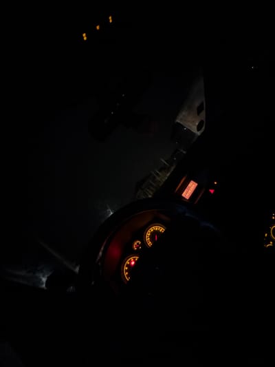 The image displays the interior of a vehicle at night, situated in the city of Karadjovo, Bulgaria. The dashboard is prominently featured, with its gauges illuminated in orange and red hues, indicating a speedometer, tachometer, fuel level, and engine temperature. A digital display, also illuminated in orange, presents the time as "23:47", a temperature reading of "19.0", and the date as "06.06.2026". A section of the steering wheel is visible in the lower left portion of the frame. The view through the windshield and the vehicle's left side is predominantly dark, showing four distinct, faint, square orange lights in the upper left quadrant, likely external light sources. No individuals are visible within the frame.