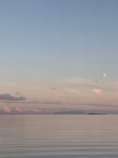 This is a landscape photograph taken at Trælnes, Norway, likely during sunset or sunrise, as indicated by the soft, diffused light and the presence of the moon. The scene is divided vertically, with the left side dominated by a pale blue sky and wispy clouds that take on a pinkish hue near the horizon. A crescent moon is visible in the upper left quadrant of the sky.

The right side of the image depicts a calm body of water, possibly a fjord or the sea, with a gently rippled surface that reflects the sky. The water appears to be a muted blue-gray, with subtle variations in tone creating a striped effect due to the light. A faint shoreline or landmass is visible in the far distance, characterized by a dark, slightly irregular shape that could be mountains or hills. There are no discernible people or man-made objects in the immediate foreground or mid-ground. The overall mood is serene and tranquil, evoked by the soft lighting and the vast, uncluttered expanse of sky and water. The time of day is suggested by the presence of both the moon and the pastel colors in the sky, indicating either dawn or dusk. The weather appears calm, with minimal wind suggested by the smooth water surface.