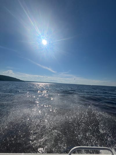 This image depicts a bright, sunny day on the water, likely a lake or sea, with a boat in motion. The sun is prominently featured, casting a bright glare and radiating light across the clear blue sky. Wispy clouds are scattered across the sky, adding texture to the scene.
The right side of the image shows the dark, choppy water being disturbed by the boat's movement, creating a spray of white water and foam. Sunlight glints off the water's surface, creating a shimmering effect. A white railing, likely part of the boat, is visible on the far right edge of the frame. In the distance, a dark, tree-covered landmass or coastline is visible along the horizon.
The overall impression is one of a dynamic and refreshing experience on the water. The bright sun and clear sky suggest pleasant weather, possibly late morning or early afternoon. The location context of Siukterka, Russia, hints at a potential setting in Northern Europe. There is no visible text in the image.