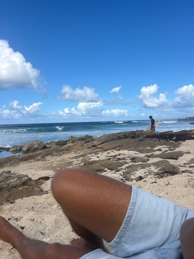 The scene depicts a coastal area in Le Moule, France, viewed from a low perspective on a sandy beach. In the immediate foreground, the lower legs and light blue denim shorts of an individual with dark skin are visible, positioned on light-colored sand mixed with small rocks and dark organic debris.
The midground features a rugged shoreline composed of dark, layered rock formations interspersed with patches of sand and seaweed. Small tide pools are present among the rocks on the left. A male individual, wearing a dark long-sleeved top (likely a wetsuit) and patterned board shorts, is walking right-to-left across the rocky beach. He carries a white surfboard under his right arm.
In the background, the ocean extends to the horizon, exhibiting moderate waves breaking towards the shore. The water appears deep blue-green. The sky above is clear and bright blue, adorned with scattered white cumulus clouds. On the distant right horizon, a faint landmass with sparse vegetation and structures is discernable. The scene is illuminated by natural daylight.