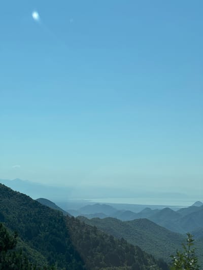 A wide-angle, slightly hazy, daytime, outdoor shot captures a view of rolling, densely forested green mountains and a distant, shimmering body of water under a bright, clear, pale blue sky. In the upper left quadrant of the sky, a small, bright, oval-shaped lens flare is visible, along with a fainter, smaller bright spot to its right. The foreground and midground are dominated by the undulating, dark green slopes of mountains covered in thick vegetation, with the nearest mountain face on the left showing a more textured, detailed coverage of trees. In the background, successive layers of mountain ranges recede into the distance, becoming progressively lighter and more muted in color due to atmospheric haze, culminating in the faint outline of a body of water that appears to be a lake or sea. The overall impression is one of natural, expansive, and serene mountain and water scenery.