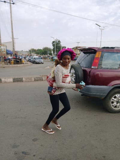The image displays a daytime street scene, with the photograph currently rotated 90 degrees clockwise. When viewed upright, a person stands prominently in the foreground on the right side of a paved road.
The person has dark skin and is wearing a vibrant pink, satin-like bonnet or head covering over what appears to be dark hair. They are dressed in a long-sleeved top with horizontal black and white stripes. Below this, a garment with a red, white, and blue pattern is visible, possibly a skirt or a fabric tied around the waist. They wear form-fitting black leggings and simple white flip-flops. The person is holding a light blue smartphone in their right hand, positioned in front of them, and is looking towards the camera with a slight smile. Their body is angled slightly, with their left arm bent and hand not visible.
Behind the person, on the right side of the frame, a maroon or dark red SUV is partially visible, parked on the side of the road. Its rear section, including a spare tire mounted on the back, and one of its wheels, are clearly seen.
The road surface in the foreground and mid-ground is asphalt, appearing somewhat worn with a few lighter patches. To the left of the road, in the mid-ground and background, there is a street extending into the distance. Along this street, various low-rise buildings, possibly commercial establishments or market stalls, are visible. There are also several utility poles with power lines crisscrossing the sky. Sparse trees with light green foliage are scattered throughout the background. Other vehicles can be faintly discerned further down the street, suggesting an active urban or semi-urban environment.
The sky above is overcast or hazy, indicating a bright but possibly not sunny day. No discernible text is visible anywhere in the image. The overall impression is that of a casual moment captured on a street.