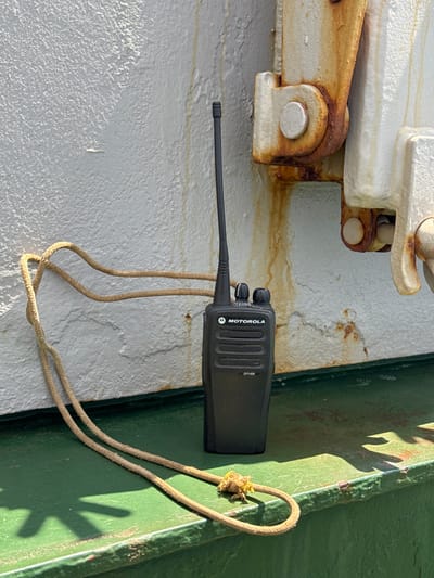 This media file shows a close-up, eye-level view of a black Motorola DP1400 walkie-talkie attached to a green metal surface with a tan lanyard. The walkie-talkie is oriented horizontally, with its antenna extending to the left. The lanyard is looped and attached to the walkie-talkie's clip at the top.
The background consists of a white, textured surface on the left, possibly a wall or bulkhead, with rusty metal fixtures and stains. To the right of the walkie-talkie is the green metal surface, which appears to be part of a ship or industrial structure. Shadows cast by an unseen object are visible on the green surface, suggesting it is daytime and sunny. There is also a thin, horizontal line of light or reflection running along the top edge of the green surface. The overall scene has a weathered, industrial feel.
The walkie-talkie has the "MOTOROLA" logo and "DP1400" text visible. There are two black knobs on the top of the device. The lanyard is made of woven rope and appears slightly frayed. The setting suggests an outdoor environment, likely on a vessel or in a port, given the weathered materials and the style of the metalwork. There are no people visible. The lighting indicates it is likely late morning or early afternoon, with strong sunlight. The rust and stains suggest exposure to the elements.