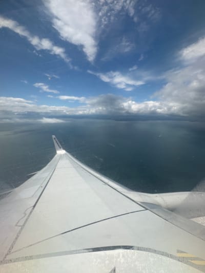 An aerial view from an airplane window shows a vast expanse of dark blue water below, with faint lines or patterns visible on the surface. A portion of the airplane's white wing, with the "RYANAIR" logo visible on the wingtip, extends into the foreground from the left. The sky above is a mix of bright blue and dramatic, heavy grey clouds, with streaks of white clouds scattered across the horizon. The overall impression is one of being in flight over a large body of water under a changeable sky. The location is not identifiable as a specific place in Unknown, Ireland from the visual information provided.