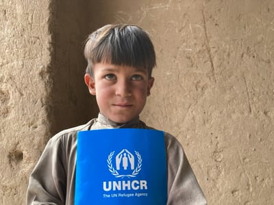 The image shows a young boy, approximately 10 years old, standing in front of a mud-brick wall. He is wearing a brown tunic and holding a blue pamphlet in front of him. The pamphlet is emblazoned with the UNHCR logo and the text “The UN Refugee Agency”. The boy is looking directly at the camera with a serious expression. The coordinates provided place the scene in Saranan, Pakistan, in a rural area characterized by mud-brick dwellings and simple living conditions.
