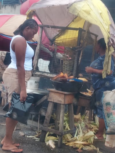 In Alagbede, Nigeria, a woman with braided hair and a white tank top stands at a market stall, holding a black plastic bag. She is wearing tan pants. Another person, wearing a blue t-shirt and jeans, is visible behind the stall. The stall is covered by a yellow and white awning and displays fried plantains and corn. Scattered corn husks are on the ground around the stall. The ground appears wet, suggesting it may have recently rained or is a damp area. The scene captures a typical street market environment in Nigeria.