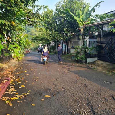 The image depicts a narrow, paved road in Kecamatan Tamansari, Indonesia, at the coordinates -6.635 latitude and 106.738 longitude. Two people are walking away from the camera on the road, one wearing a blue shirt and pants, the other wearing a white headscarf and a black jacket. A third person is driving a motorcycle towards the camera. There are trees and foliage on either side of the road.  A black gate and a house with a white facade can be seen on the right side of the image. The road is made of grey asphalt with yellow fallen leaves scattered across it.  The scene is quiet and tranquil, with the only sounds being the motorcycle engine and the rustling of leaves.