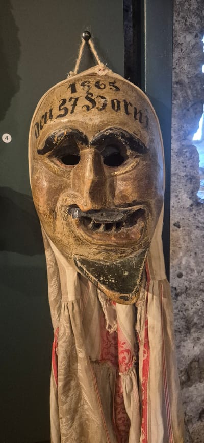 This media file depicts a close-up, rotated view of an artifact, likely a mask, displayed on a dark surface. The mask is made of wood and painted with a distressed, aged appearance. It features exaggerated facial elements, including wide-open eyes, a gaping mouth with visible teeth, and prominent cheekbones. Black paint is used for the eye sockets and mouth, with some detailing suggesting eyebrows or other facial features.
Text is visible on the left side of the mask, oriented vertically. It appears to be a date and possibly a name or identifier, reading "oct 37 SSORN". The numbers "1865" are also partially visible below this.
The mask is adorned with strings and a small, dark spherical object on the left side, suggesting it was either meant to be worn or has been prepared for display. A fabric, likely a cloth or costume element, is draped behind and around the mask. This fabric is a light tan color with repeated patterns of red stripes and possibly floral or damask designs in red and white.
The background suggests an indoor exhibition setting. The top edge of the image shows a rough, stone-like texture, possibly part of a wall or display structure, with some bright light emanating from above. The overall impression is of a historical or cultural artifact being presented for viewing. The specific location context provided is Sargans, Switzerland. There is no visible indication of time of day, weather, or specific emotions being conveyed beyond the inherent expression of the mask itself. A small, white circular tag with the number "4" inside is visible on the dark surface in the lower left corner of the frame.