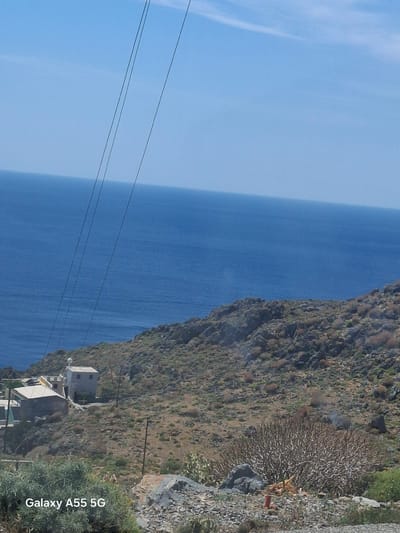 The image shows a view of the sea and a cliffside in Plakias, Greece. The left side of the image is dominated by the blue expanse of the sea, with a bright blue sky above. Three parallel electrical lines can be seen stretching across the sea and sky. On the right side of the image is a rocky cliff covered with sparse vegetation. Several buildings are nestled into the cliffside.