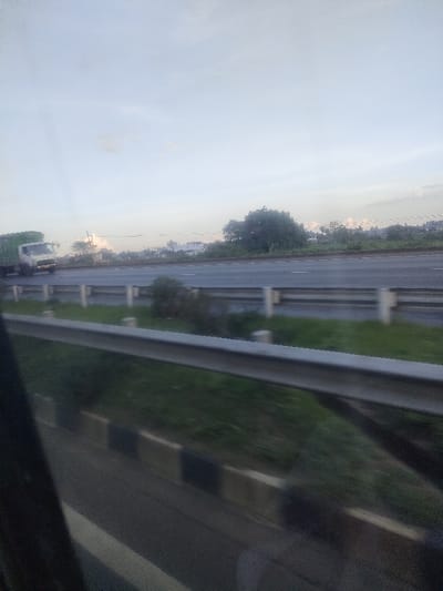 This image depicts an outdoor highway scene captured from a moving vehicle in Kalimoni ward, Kenya, during daylight hours. The foreground is partially obscured by a dark frame, likely a window, through which a silver metal guardrail is visible, supported by light-colored concrete posts. Beyond the guardrail, a grassy median strip separates the viewer's immediate perspective from a multi-lane paved road. A white truck with a green cargo container is present in the far-left lane of the visible highway, moving from left to right. The background features extensive green foliage and trees, with several light-colored buildings and structures discernible in the distance. The sky above is light blue with scattered white clouds. No individuals are visible in the scene.