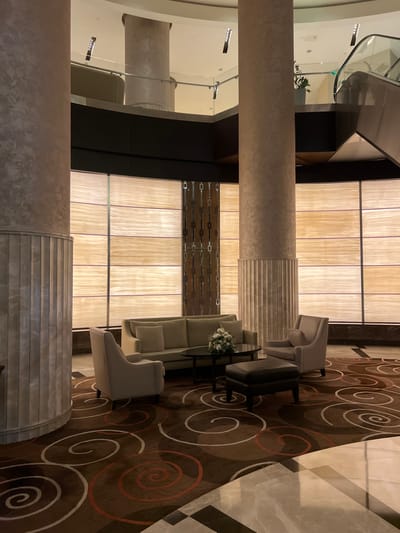 This image depicts a modern, multi-story lobby area within a building in Bağcılar, Turkey. The scene is characterized by tall, cylindrical structural columns with a textured, light beige finish, featuring fluted decorative bases. A mezzanine or upper level is visible, encircled by a glass and metal railing, with an escalator ascending on the right side.
The background wall features a series of large, horizontally oriented, backlit translucent panels, likely made of onyx or a similar material, providing ambient illumination. A central vertical decorative panel with a dark, metallic, chain-link-like design separates segments of these illuminated panels.
In the mid-ground, a lounge seating arrangement is positioned on a brown carpet adorned with a repeating pattern of large white, red, and brown spirals. The furniture includes a light beige three-seater sofa with multiple pillows, flanked by two matching single armchairs. A dark-colored round coffee table, holding a white floral arrangement, is placed in front of the sofa. A dark brown or black leather ottoman is situated in the foreground of the seating area.
The flooring in the immediate foreground transitions from the spiral-patterned carpet to a geometric pattern of polished black and white tiles. Overhead lighting is recessed. No individuals are present in the depicted scene.