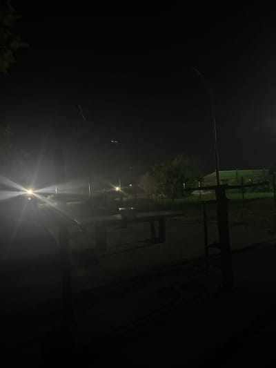 The image depicts a nighttime outdoor scene, possibly a park or recreational area, in Egorlykskaya, Russia. The foreground is dominated by dark shadows, obscuring much of the immediate surroundings. In the mid-ground, several dark, rectangular table-like structures are visible, suggesting picnic tables or benches. These are situated on a paved surface, possibly brick or concrete, with a distinct pattern.
To the left, a bright light source, likely headlights of a vehicle, casts a strong glare and lens flare across the scene, illuminating the air with a hazy glow. Several streetlights are present throughout the mid-ground and background, casting pools of light and contributing to the overall low-light atmosphere.
In the background, a fence runs horizontally, separating an area with some vegetation and trees from the immediate foreground. Beyond the fence, a dimly lit building or structure with a green roof is visible. The sky is dark, with only a few faint stars or distant lights visible. The overall impression is one of an empty, dimly lit public space at night.