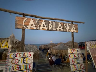 The image depicts a beachside establishment in Toroni Municipal Unit, Greece, at dusk. A weathered wooden sign with "CASABLANCA" painted in white letters against a distressed wood background hangs from a rustic wooden frame, identifying the location. In the foreground, several smaller signs list offerings such as "COCKTAILS", "COFFEE", "FRESH JUICE", "BEERS", and "SNACKS", painted in colorful block letters on white, distressed wooden planks. Behind these signs, thatched umbrella-shaped roofs provide shade over seating areas on the sandy beach. A few people are seated under the umbrellas, some appearing to be patrons. The sea is visible in the background under a clear, pale blue sky, with a hint of a sunset or moonrise indicated by a faint orange glow on the horizon. The overall atmosphere suggests a relaxed, casual beach bar setting.