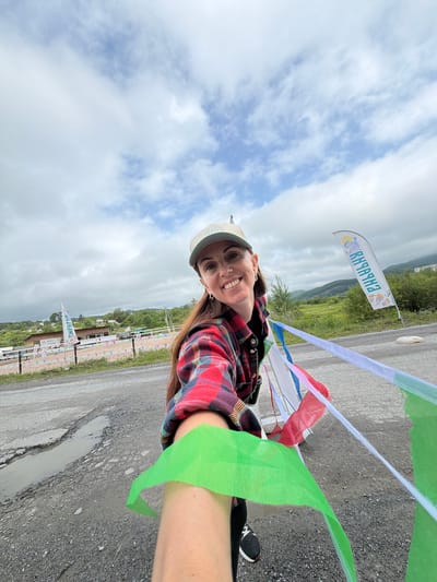 This is a selfie of a woman with long brown hair, wearing a red and black plaid shirt, a light-colored baseball cap, and a green streamer draped over her arm. She is smiling and looking directly at the camera. The photo is taken outdoors with a cloudy sky.
In the background, there is a road with puddles and cracks, suggesting it might have recently rained. There are also several flags and banners visible, some with colorful streamers, which could indicate an event or a race. One flag has text on it that appears to be in Cyrillic, reading "КЫЗЫЛ-ХАЙЯ". The surrounding landscape shows rolling green hills and trees.
The overall impression is of a cheerful moment captured during an outdoor activity, possibly a race or a festival, given the festive decorations and the woman's energetic pose. The weather appears overcast but not actively raining. The location context provided is Teploozersk, Russia.