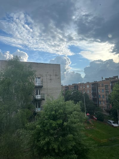 The image shows an upward view in a city, likely Dmitrievskaya, Russia. The sky is dominated by a mix of cumulus clouds and patches of blue. Two brick buildings are visible, one a taller apartment building with multiple windows and the other a lower building. Trees with green foliage are positioned in the foreground, partially obscuring the lower building and the apartment building. A playground with red and yellow structures is visible in the space between the buildings and the trees. A car is parked in the distance.