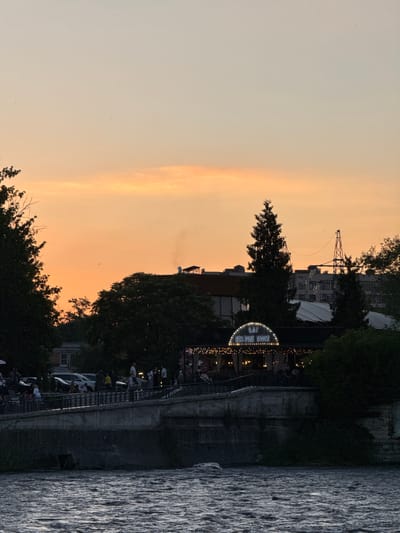This media file captures a sunset scene at what appears to be a waterfront restaurant or entertainment venue, possibly located in Karaman 1-YY, Russia. The sky is painted with hues of orange and yellow, indicative of dusk. In the background, several buildings and a tall industrial-looking structure with a crane are visible.
The main focus of the scene, illuminated by string lights, is a semi-circular structure with what seems to be a sign that reads "SAT PIZZA ROCK" in neon letters. People are gathered on a walkway or patio area in front of this structure, enjoying the evening. Some are seated, while others appear to be standing and socializing. The presence of the water to the right of the scene suggests a river, lake, or canal. The water's surface is textured, reflecting the dimming light. The overall atmosphere appears relaxed and festive, with people enjoying an evening out. The time of day is clearly sunset, with the warm glow in the sky and the artificial lights starting to dominate the foreground.