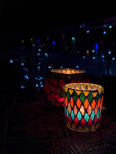 Two cylindrical candle holders with mosaic patterns are mounted on a wall. The front candle holder has a pattern of yellow, green, orange, light blue, and red diamonds. The second candle holder has a red diamond pattern. The wall has a dark, patterned surface, possibly tile with a floral motif. In the background, out-of-focus lights of various colors suggest a festive or decorative atmosphere. The location is likely within Vinaròs, Spain.