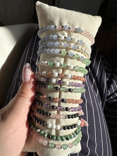 A person is holding a small, rectangular, off-white, textured pillow in their left hand. Resting on the pillow are ten beaded bracelets, all of different colors. The bracelets are arranged in a row, with each bracelet displaying a unique combination of colored beads and small silver-colored spacers. From left to right, the bracelet colors are pink, light blue, white, light green, green and white, peach and purple, green, peach and black, purple and black, and a final bracelet of green, white, and pink beads.
The person has fingernails painted with a light pink base and dark pink or purple spots. The person is wearing a navy and white striped garment.
The lighting in the scene appears bright and direct. The background is out of focus. There is no indication of this image being taken in Galiche, Bulgaria.