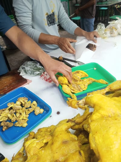 The image depicts a food preparation area within the city of Houaythat, Laos. Three individuals are visible. In the foreground, a person wearing a dark blue shirt extends their right forearm towards a stack of green plastic trays. Adjacent to them, another individual in a light blue long-sleeved shirt, featuring yellow script on the chest, uses both hands to wrap dark, cooked meat in white paper. This wrapping occurs on a white surface, likely a table or counter.
On the table in the immediate foreground, a pile of whole, cooked yellow chickens is present. To the left, a blue rectangular plastic tray contains numerous yellow, cooked chicken gizzards or similar offal. A stack of green plastic trays sits centrally, with the top tray holding yellow, butchered chicken pieces; a hand is seen selecting items from this tray. Other objects on the white surface include a pair of dark tongs, a clear plastic bag containing green leafy items, a red and white checkered cloth, and a wooden cutting board.
In the background, a third person, dressed in a dark shirt and blue jeans, stands behind a dark-framed table with bright green stools. Additional clear plastic bags, possibly containing food, are scattered on the back tables. The floor appears to be concrete. The scene suggests a bustling market stall or informal eatery setting.