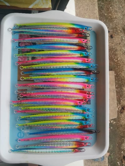 The image shows a white plastic tray filled with 21 fishing lures. The lures are arranged vertically in a row. They are all made of metal and have a silver base with colorful stripes of pink, blue, yellow, and green. Each lure has a small, sharp hook at the end and a loop at the other end. The lures are arranged in a neat, organized fashion and appear to be new and unused. The background is a blurred out view of a tiled floor. The image was taken in Kebumen, Indonesia at the coordinates -7.662, 109.647.  The image was taken indoors and shows a close-up of the tray.